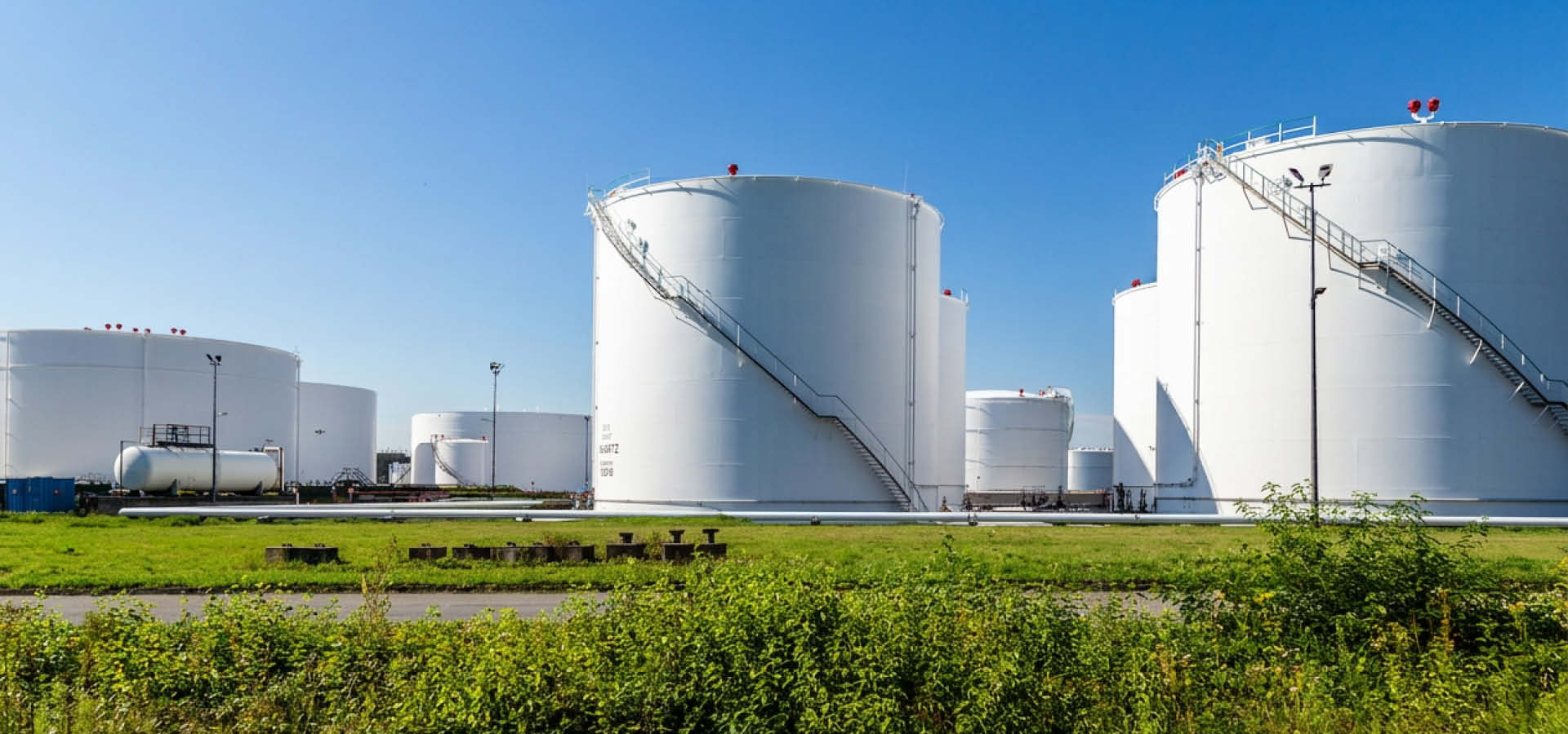 Vy över tankgård med flera vita cisterner för bränsle/petroleumprodukter under klarblå himmel